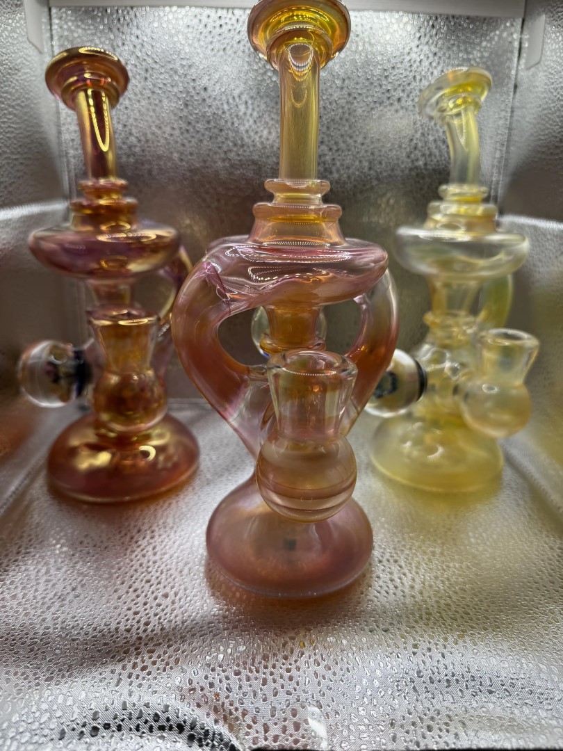 Three intricately designed glass water pipes in pink, orange, and yellow hues are displayed upright inside a metallic, condensation-lined enclosure from our mobile smoke shop in Winter Haven, Florida.
